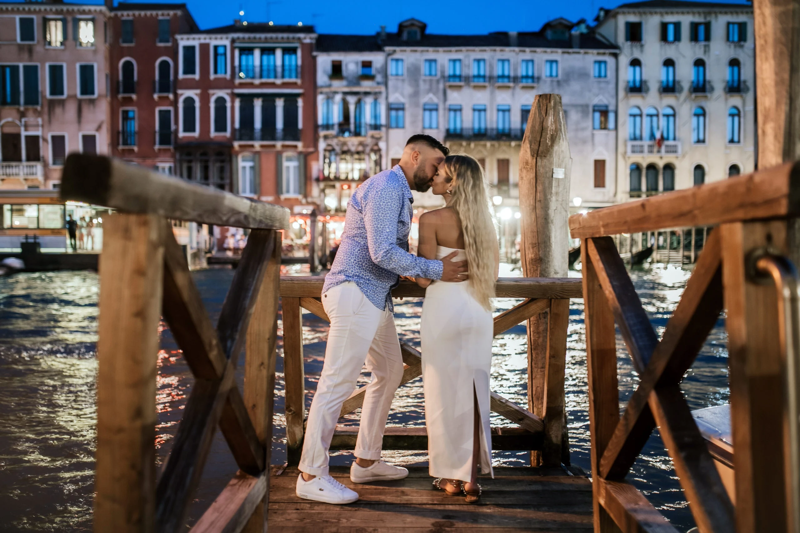Couple standing by the wooden posts as the lights come on along the canal