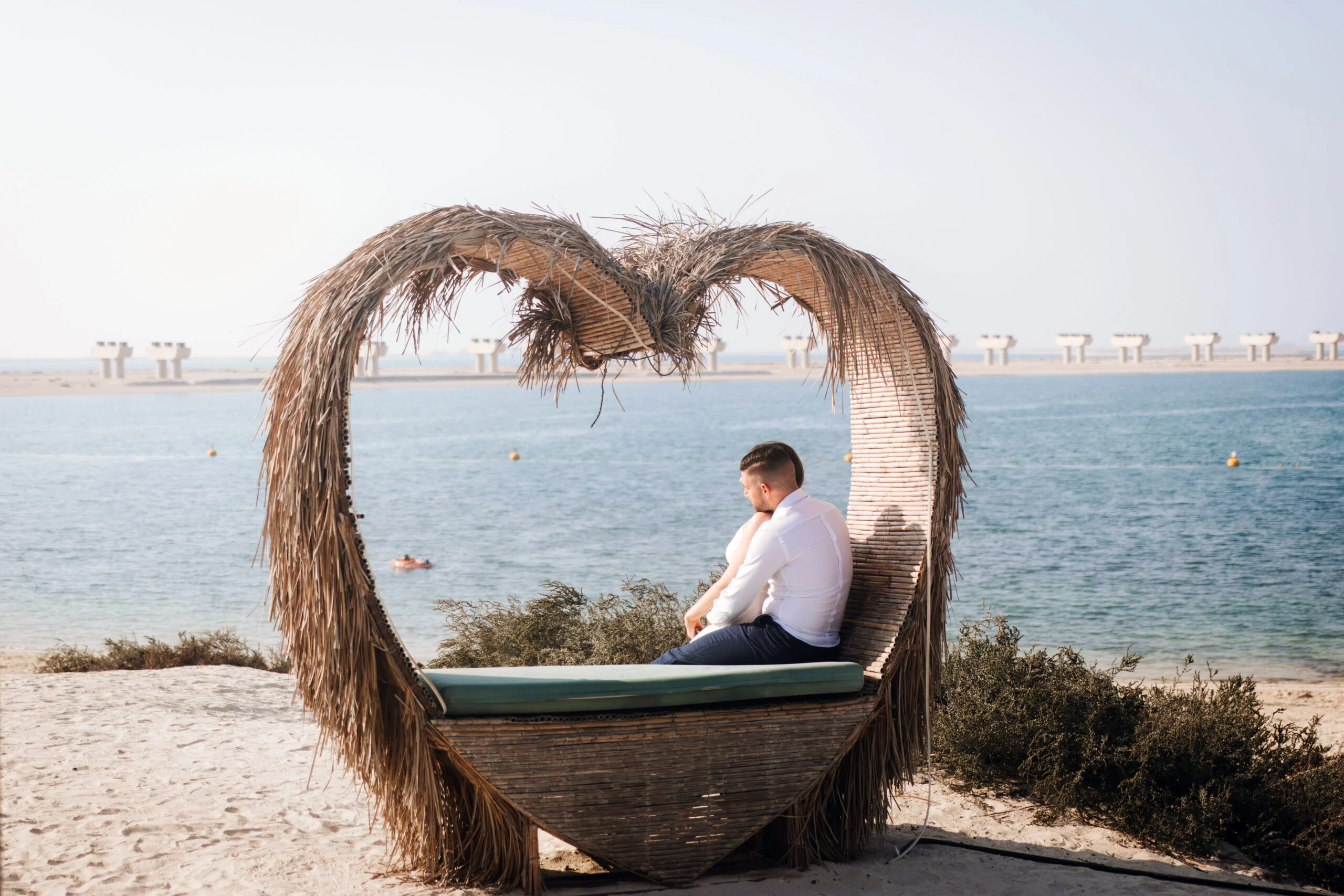 Man sitting in a woven heart-shaped beach shelter looking out to sea