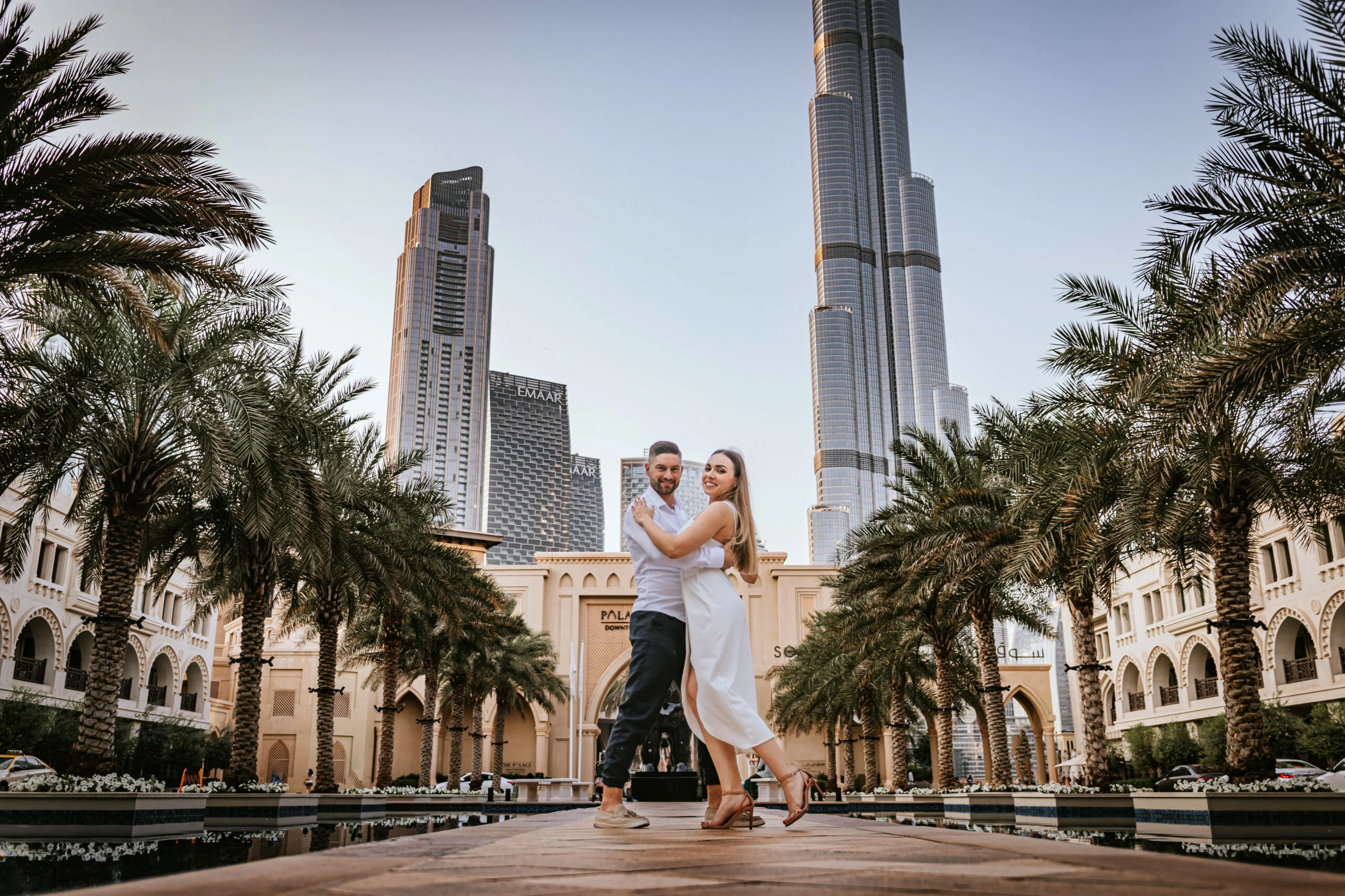 Couple standing in a palm-lined courtyard with the city towers behind