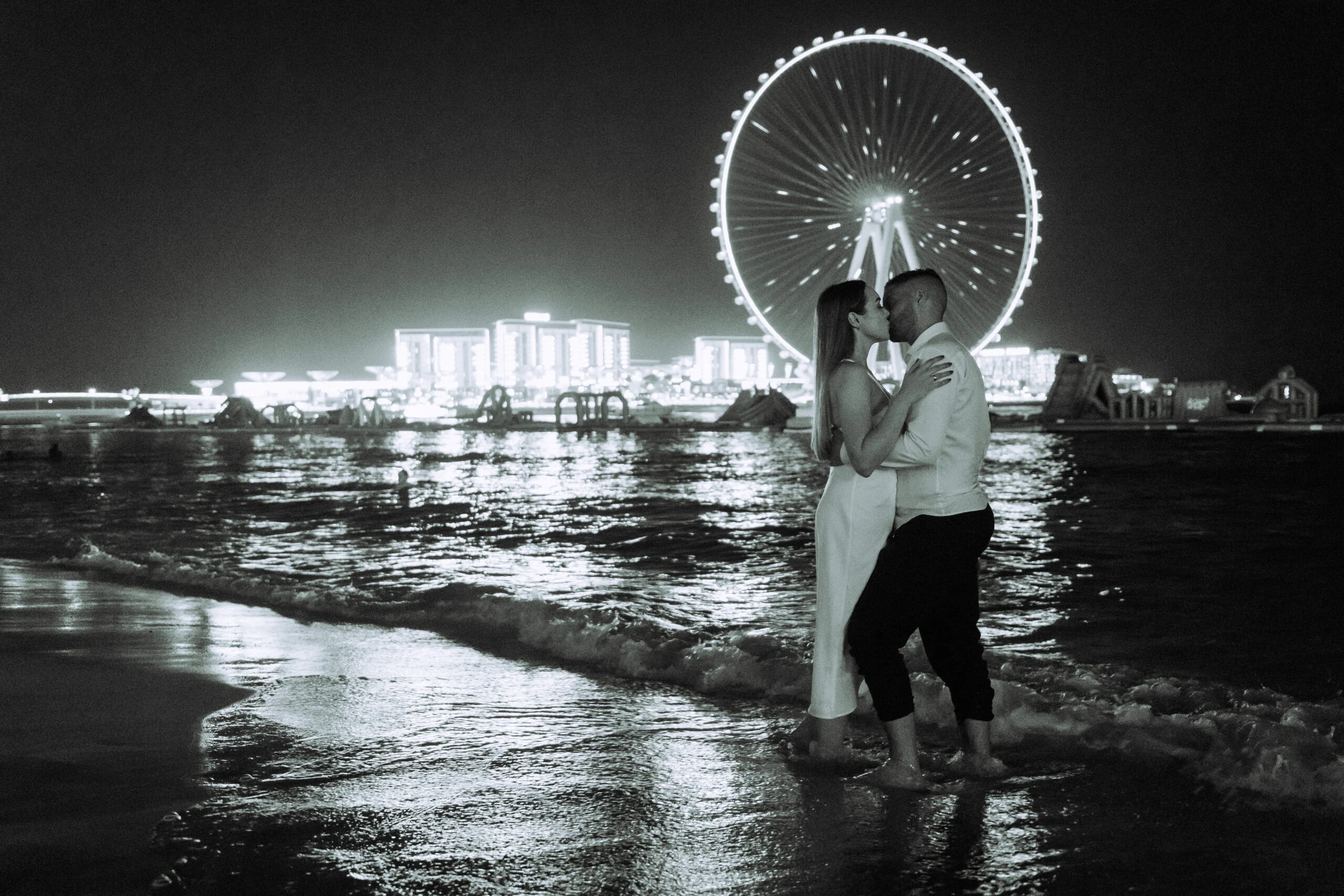 Black and white photograph of the couple on the beach with an observation wheel behind them