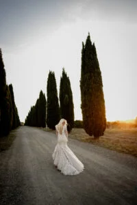 Bride walking alone up a cypress-lined gravel road at sunset
