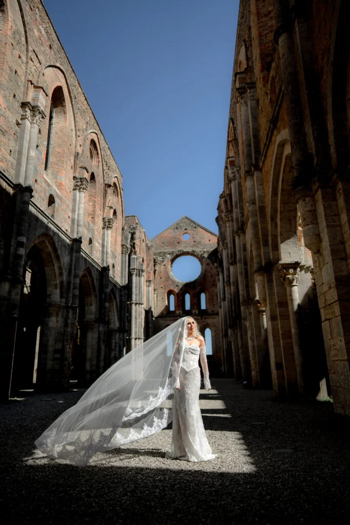 Bride standing in the nave of a roofless abbey in Tuscany, her long veil across the stone floor