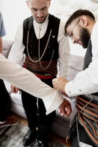 Two men in traditional Greek dress helping the groom into his shirt