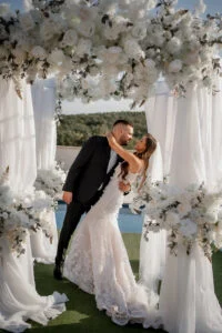 Bride and groom kissing under a flower-covered arch beside a pool at a private estate