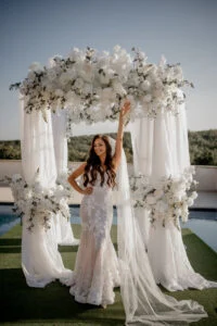 Bride holding the edge of the floral arch, her veil trailing across the grass