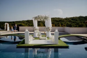 Ceremony setting on a platform above a pool, with a floral arch and white pillars