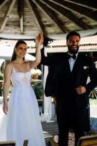 Bride and groom under a wooden pavilion, the groom raising his hand