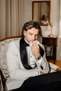 Groom in a white dinner jacket in a hotel suite, the bride reflected in the mirror behind him