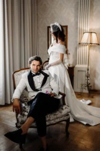 Bride standing behind the groom, who is seated in an armchair in a hotel suite