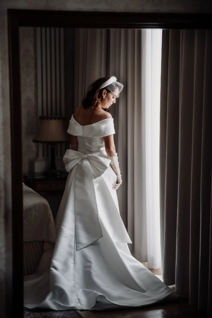 Bride in a satin gown and short veil standing by a window in a hotel room