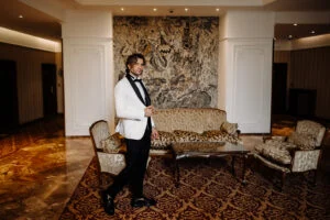 Groom in a white dinner jacket standing in a hotel lounge with a large mosaic on the wall