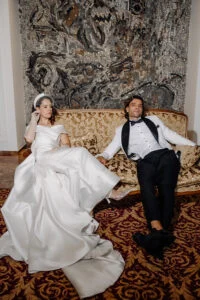 Bride and groom sitting on an antique sofa in a hotel suite, her dress spread across the floor
