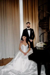 Bride seated in her gown beside a grand piano, the groom standing behind her
