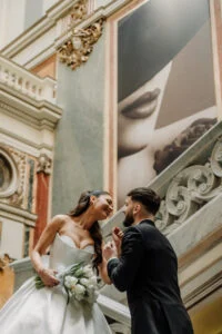 Groom leaning towards the laughing bride, who holds a white bouquet, on the gallery of a historic building