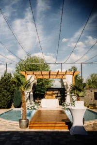 Ceremony setting beside a pool, with a wooden pergola, white drapes and flowers