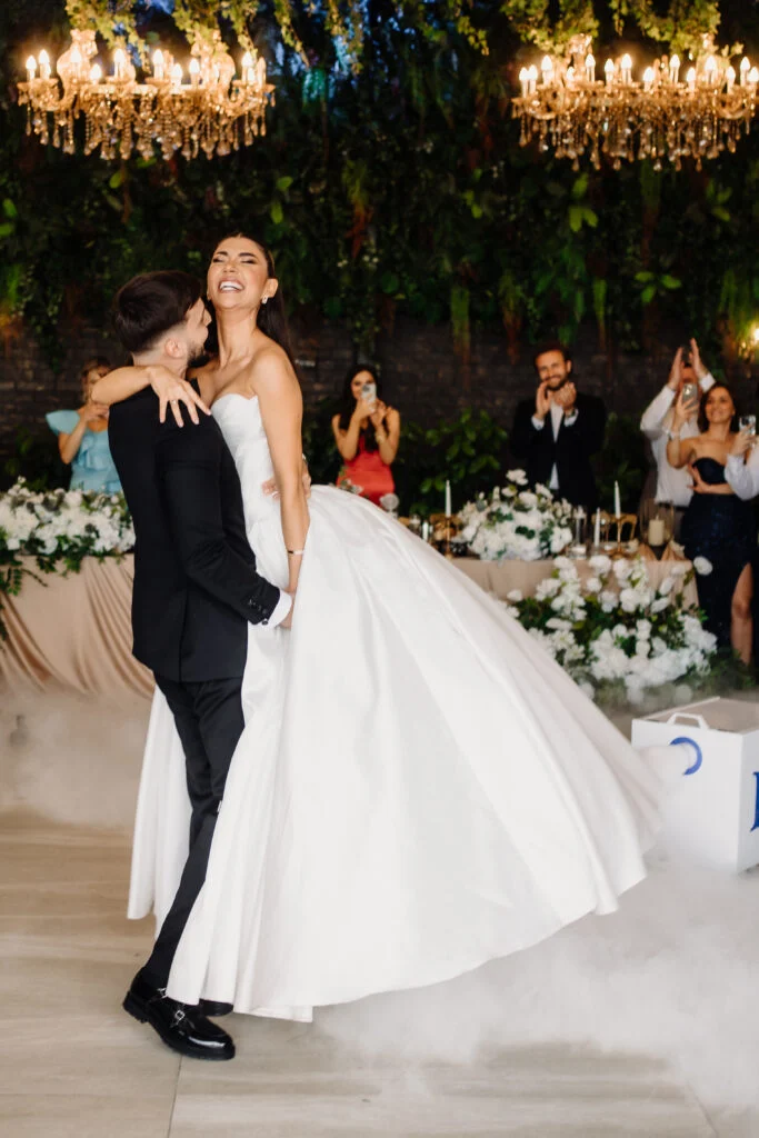 Groom lifting the bride on the dance floor beneath chandeliers, guests watching behind them
