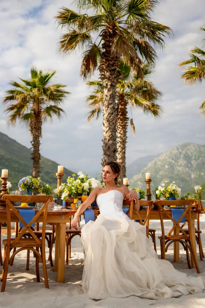 Bride seated at a long table set for the wedding dinner between palm trees