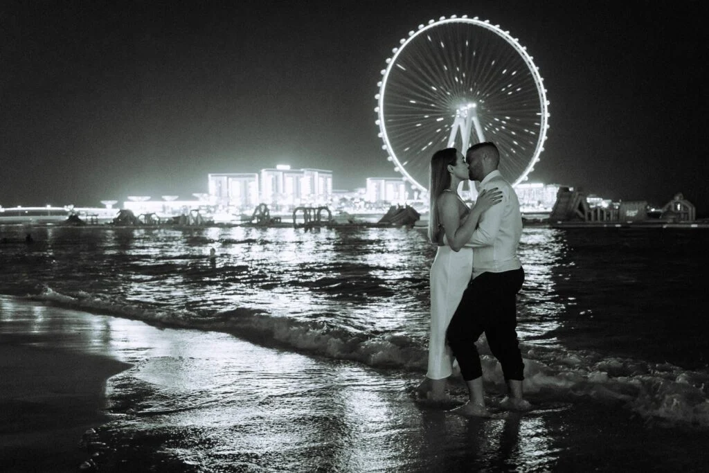 Black and white photograph of a couple in shallow water, a lit ferris wheel behind them