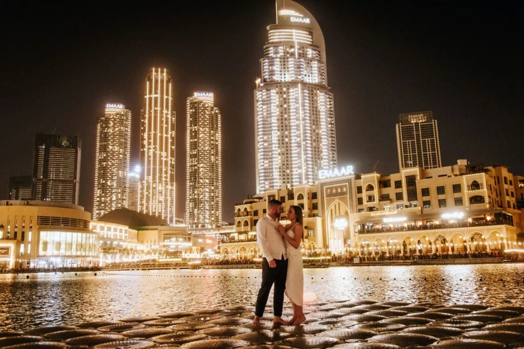 Couple embracing at the water's edge with the lit city behind them