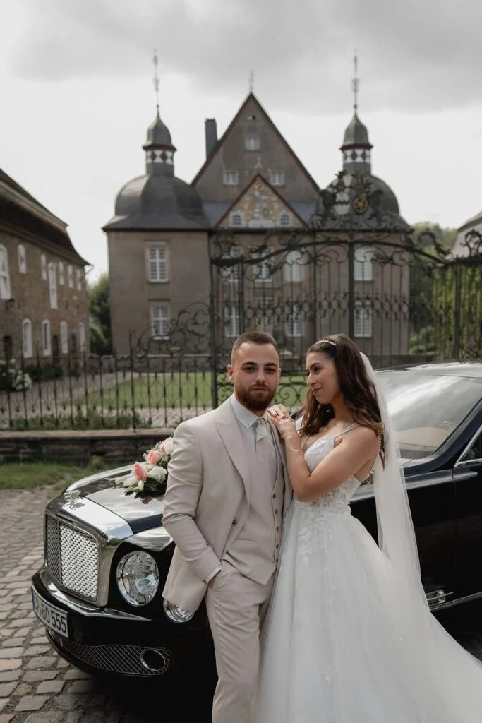 Bride and groom leaning against a black car, the castle behind them
