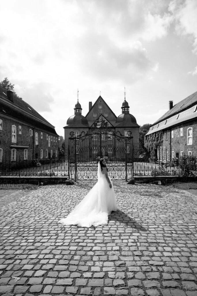 Black and white photograph of the bride on the cobbled courtyard, train spread, castle behind