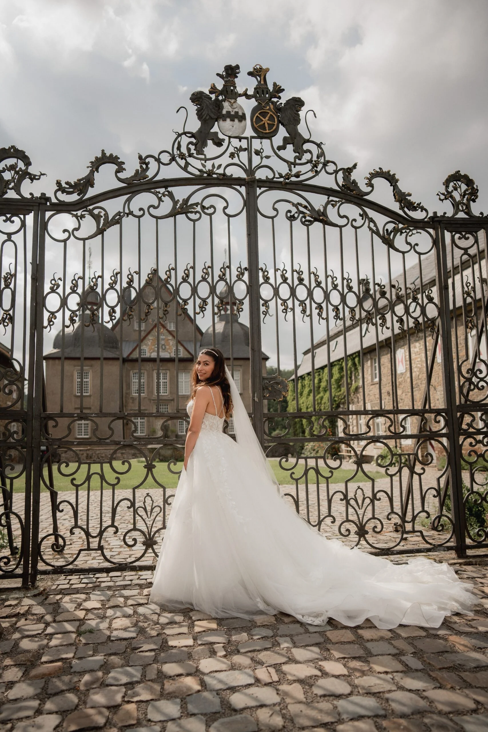 Bride seen from behind at the wrought iron castle gate, her train spread across the cobblestones