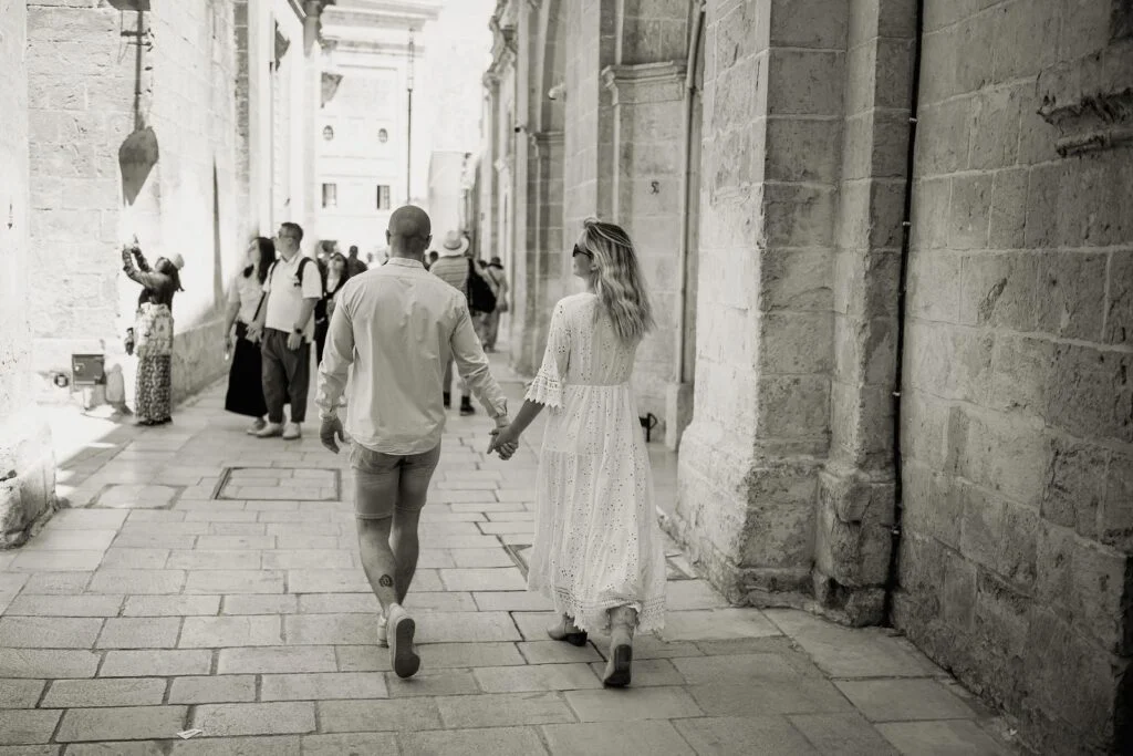 Black and white photograph of a couple walking hand in hand down a narrow street in Mdina