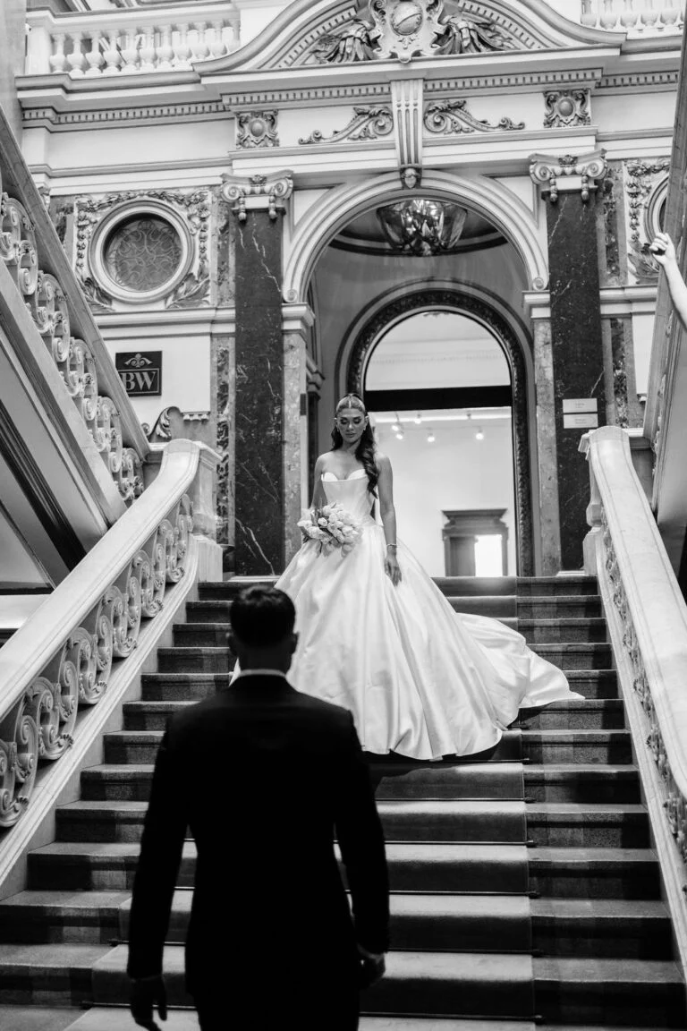 Black and white photograph of the groom watching the bride climb the staircase at Salon 1905