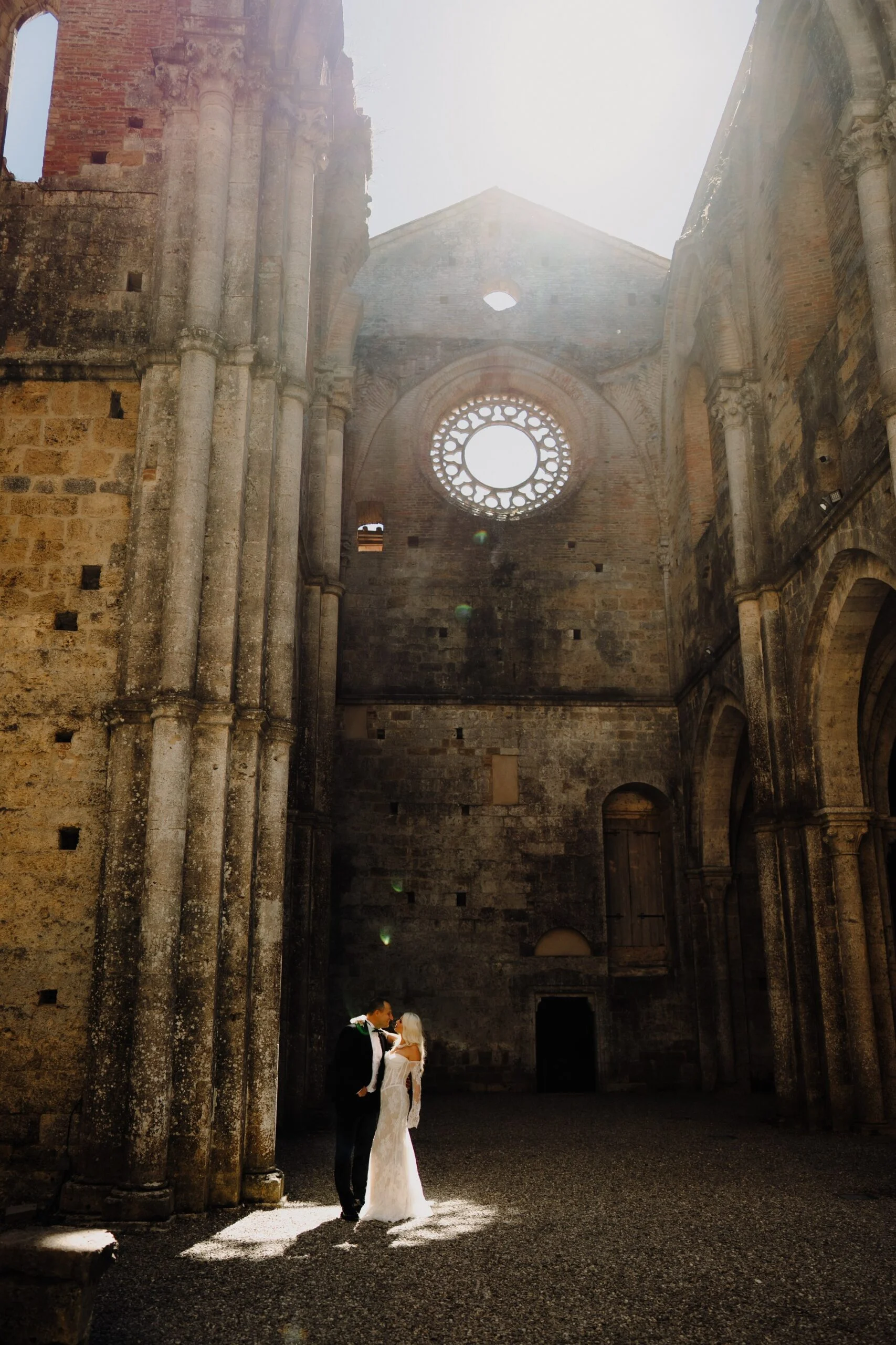 Bride and groom in a roofless abbey in Tuscany beneath the rose window