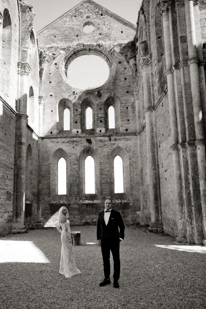 Bride and groom in the abbey, her gown lit by sunlight on the gravel floor