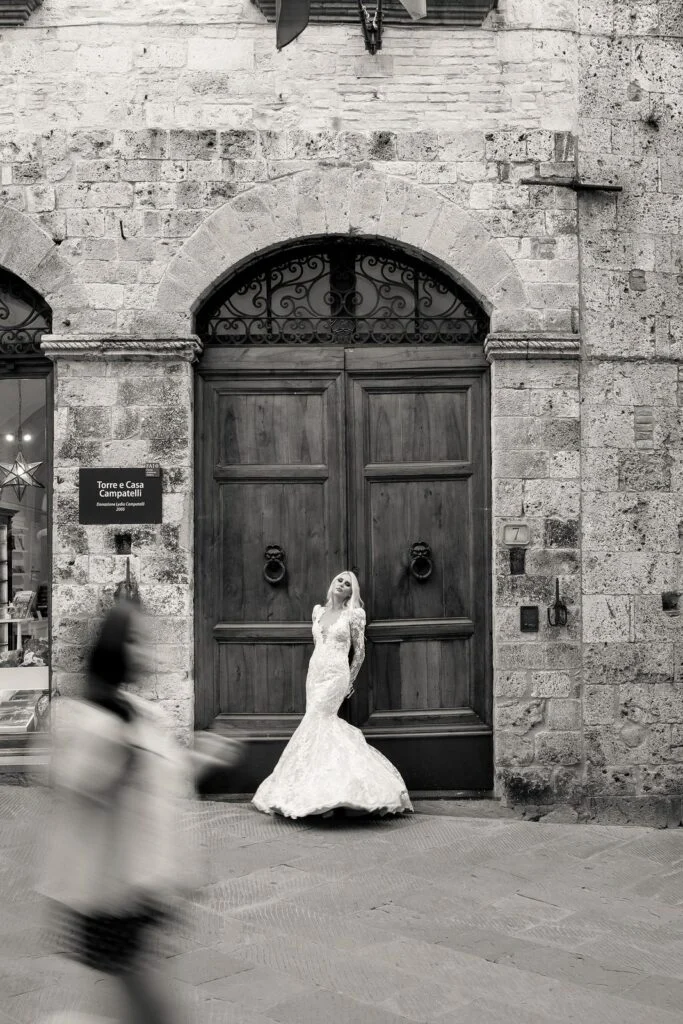 Black and white photograph of the bride before a tall wooden door, a passer-by blurred in motion